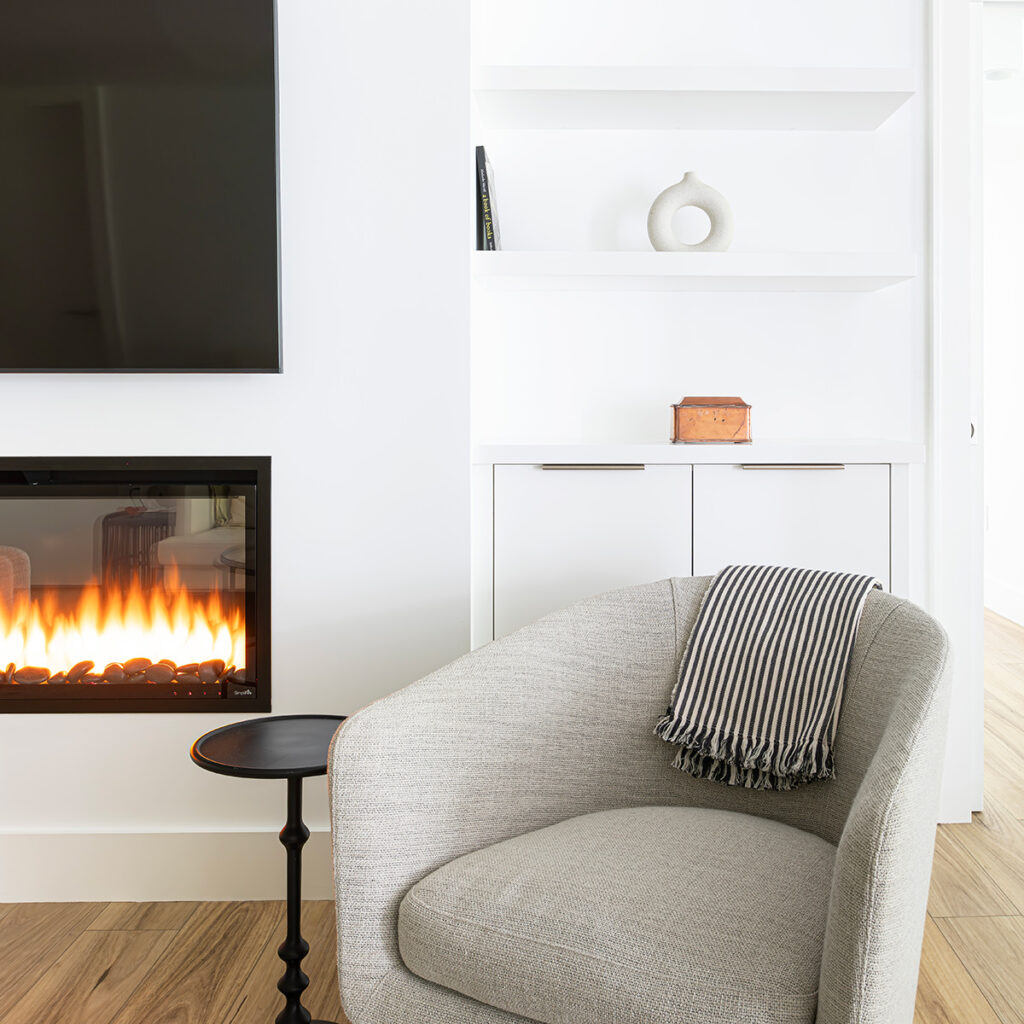 Modern living room with large TV, contemporary linear fireplace, white built in cabinets and a cozy grey cloth arm chair.