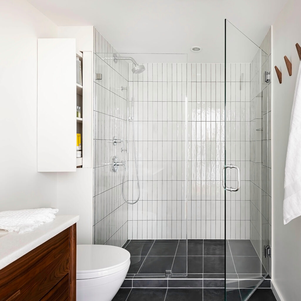 Scandinavian modern bathroom with white tiled shower, dark grey floor tile, wall storage cabinet, and mahogany vanity.